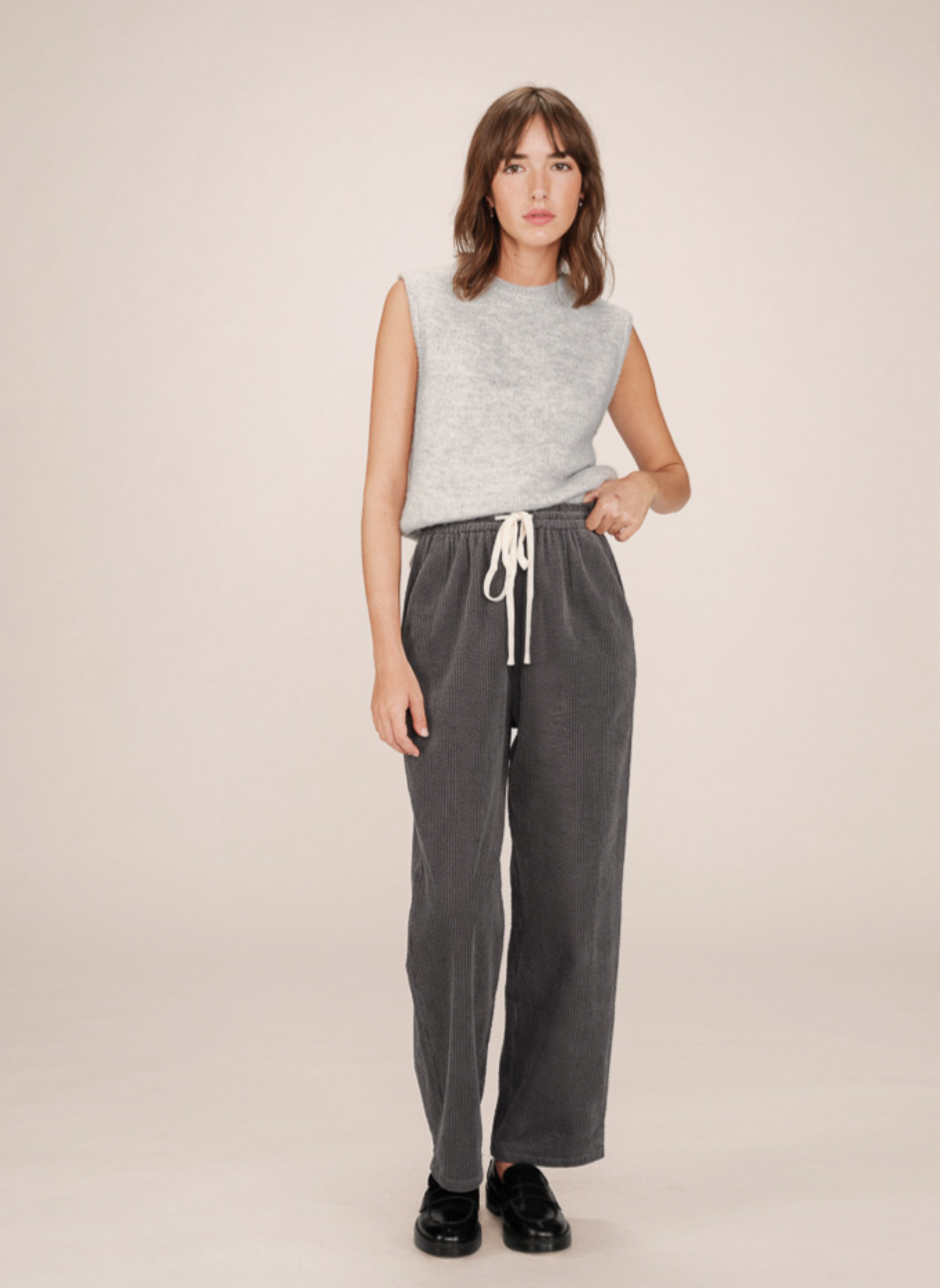 Woman wearing a sleeveless top and grey corduroy pants with cream draw string tie on a beige background
