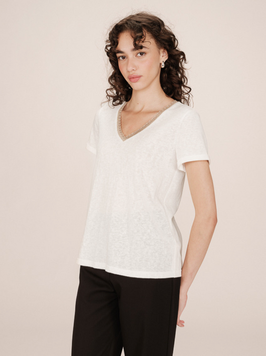 Woman wearing a white textured top with glittery gold neckline detail and black pants against a beige background