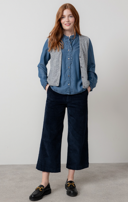 Woman wearing a blue denim shirt, gray vest, and dark blue pants against a plain background