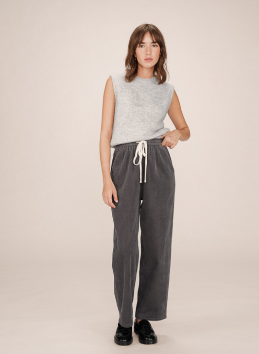Woman wearing a sleeveless top and grey corduroy pants with cream draw string tie on a beige background