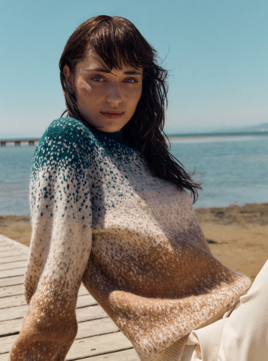 Woman wearing a patterned blue and fawn sweater sitting on a wooden dock by the water.
